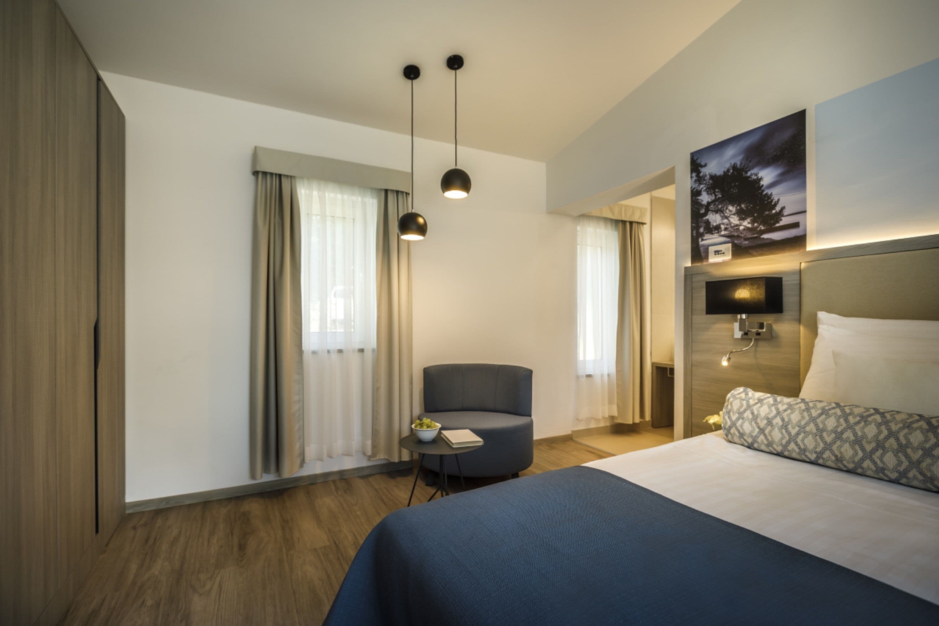 Una habitación de hotel moderna con una cama grande, una mesita redonda con un frutero, una silla y un cuadro en la pared.