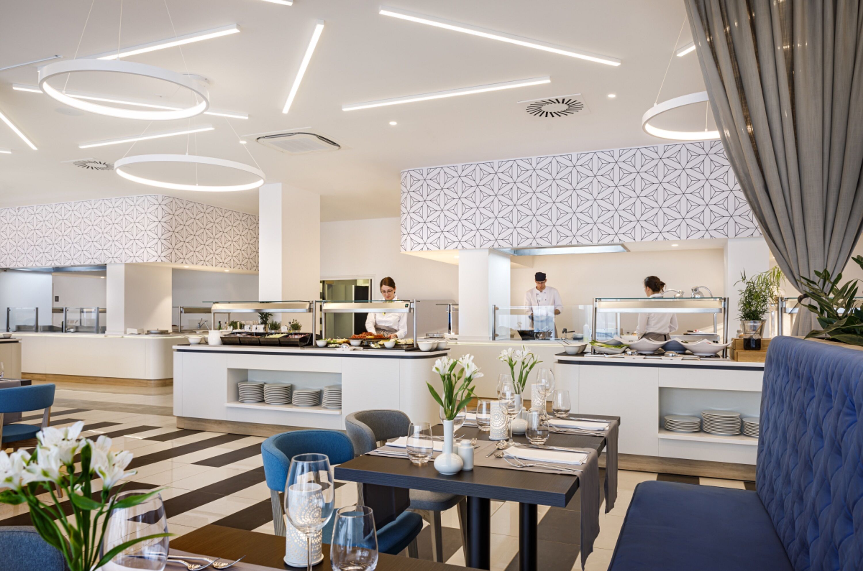 A modern hotel dining area with a buffet setup, seating arrangements, and a decorative ceiling design.