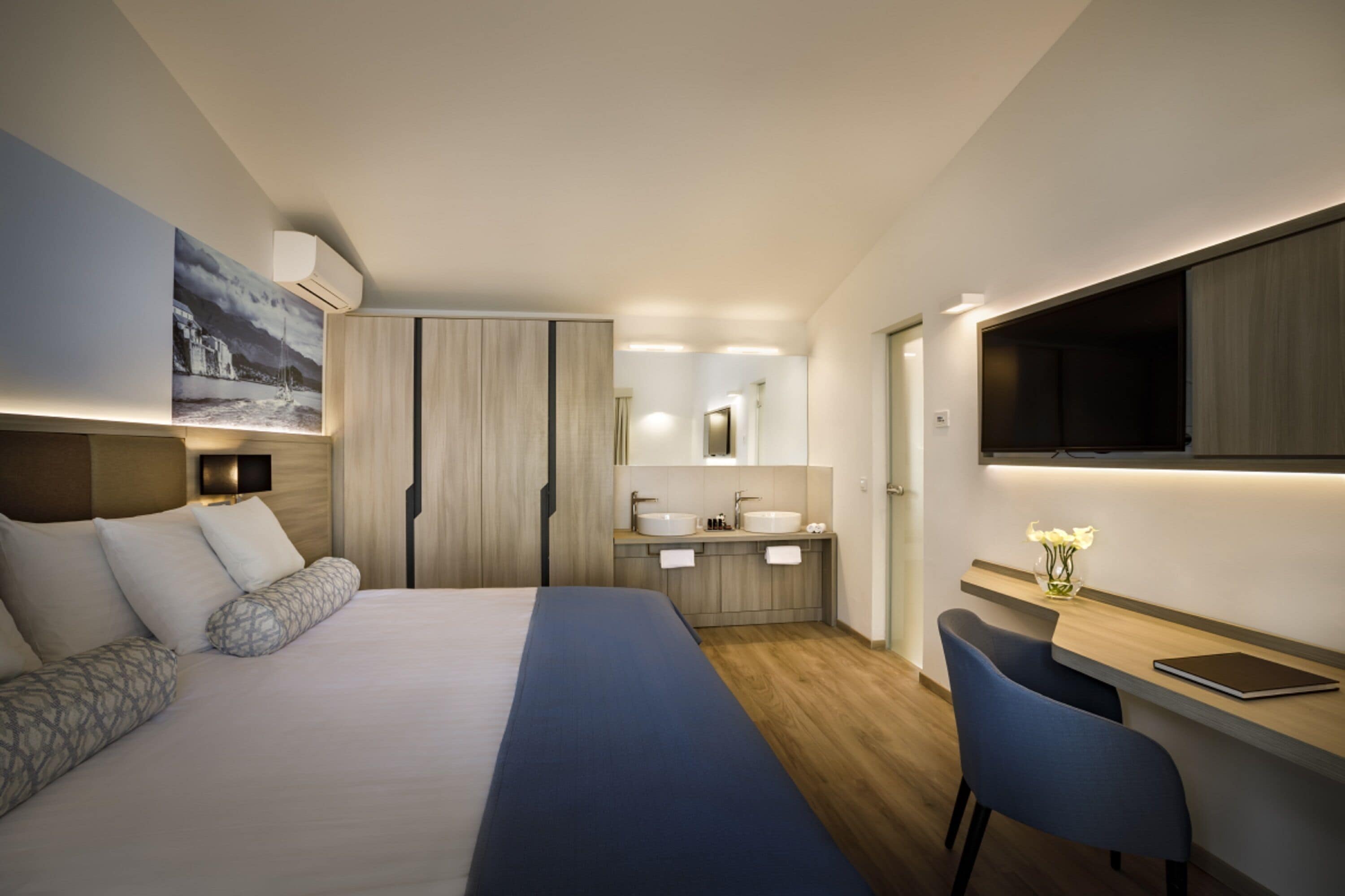 A modern hotel room with a large bed, a desk with a chair, a bathroom with a sink and mirror, and a TV mounted on the wall.