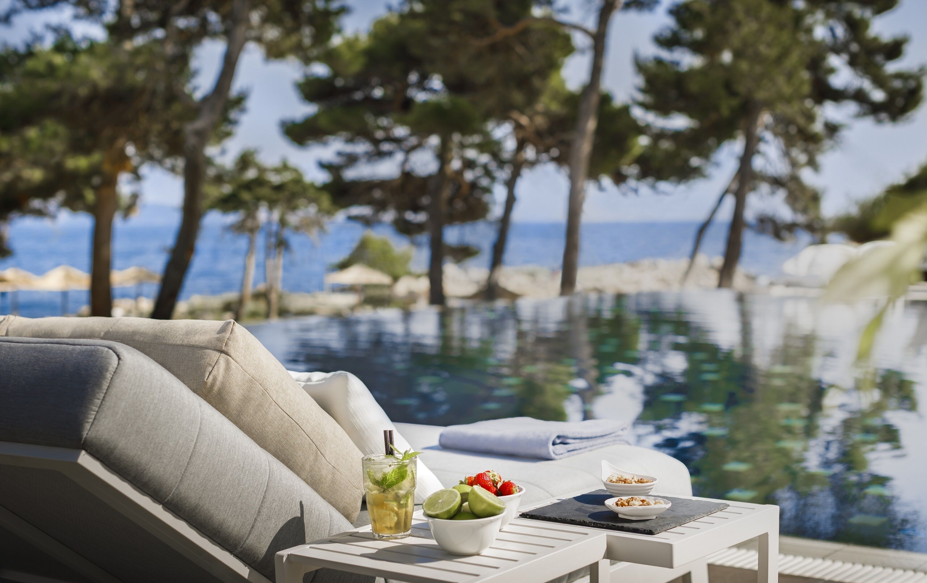 A poolside lounge area with a view of the sea, a table with a drink and snacks, and a reclining chair.