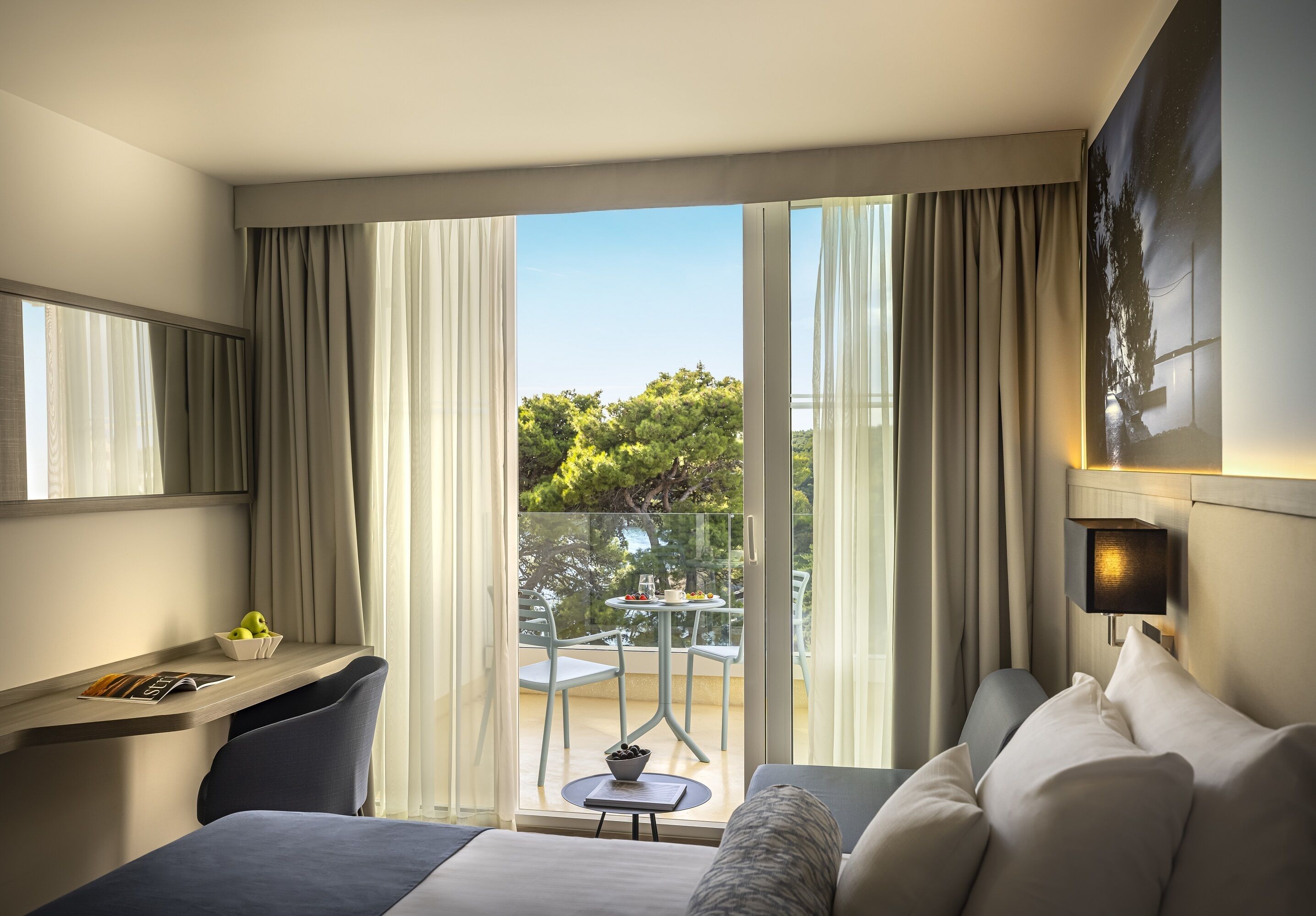 Ein Hotelzimmer mit einem Bett, einem Schreibtisch mit Stuhl, einem kleinen Tisch mit Obstschale und einem Balkon mit Tisch und Stühlen.