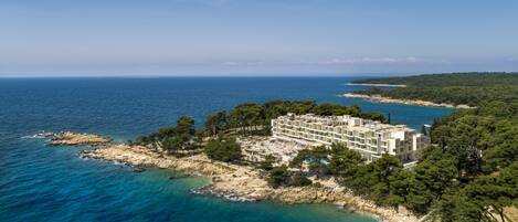 A coastal hotel with a modern design, situated on a rocky peninsula with clear blue waters and a view of the sea.