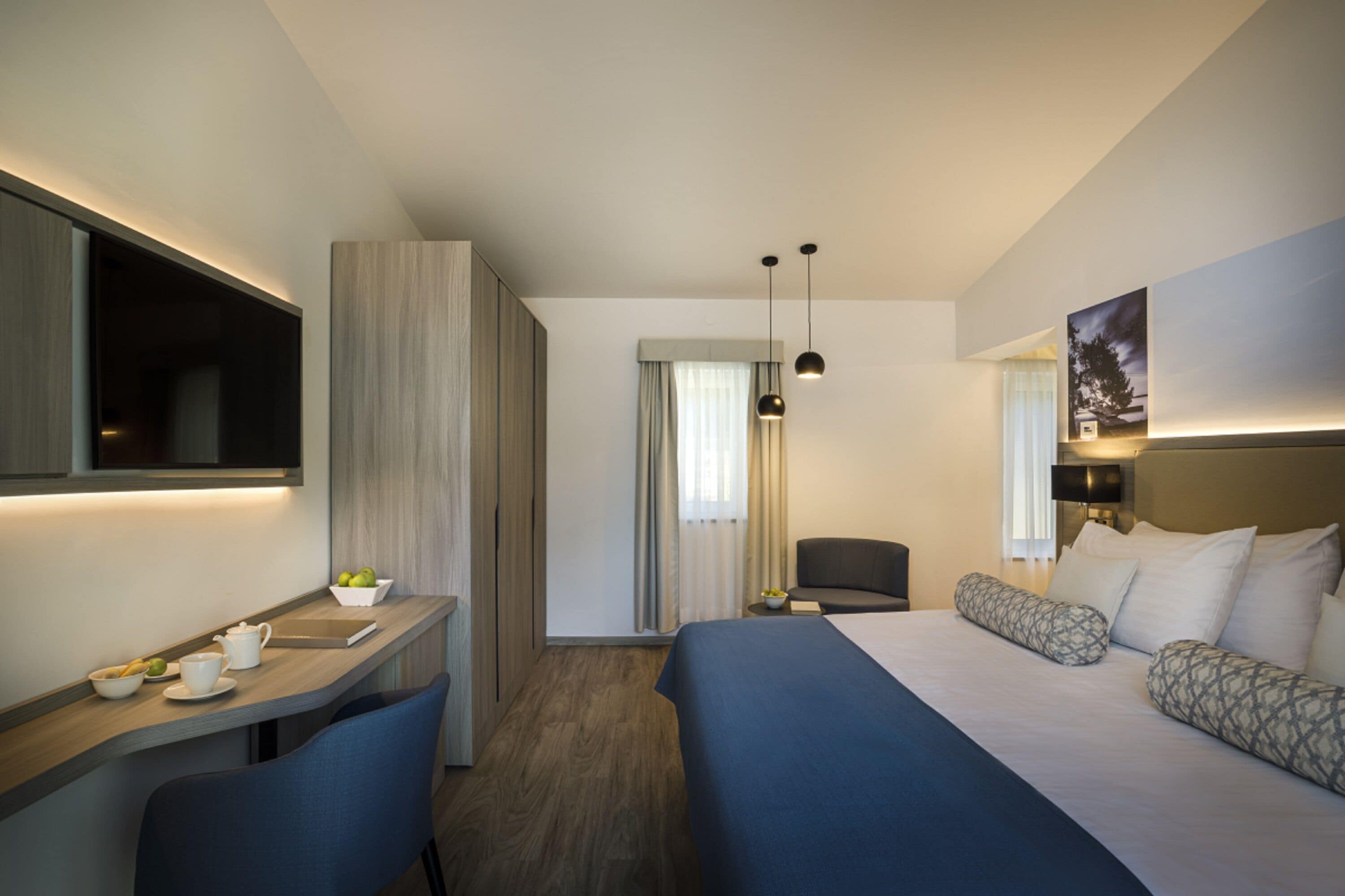 A modern hotel room with a bed, a desk with a chair, a television, and a painting on the wall.