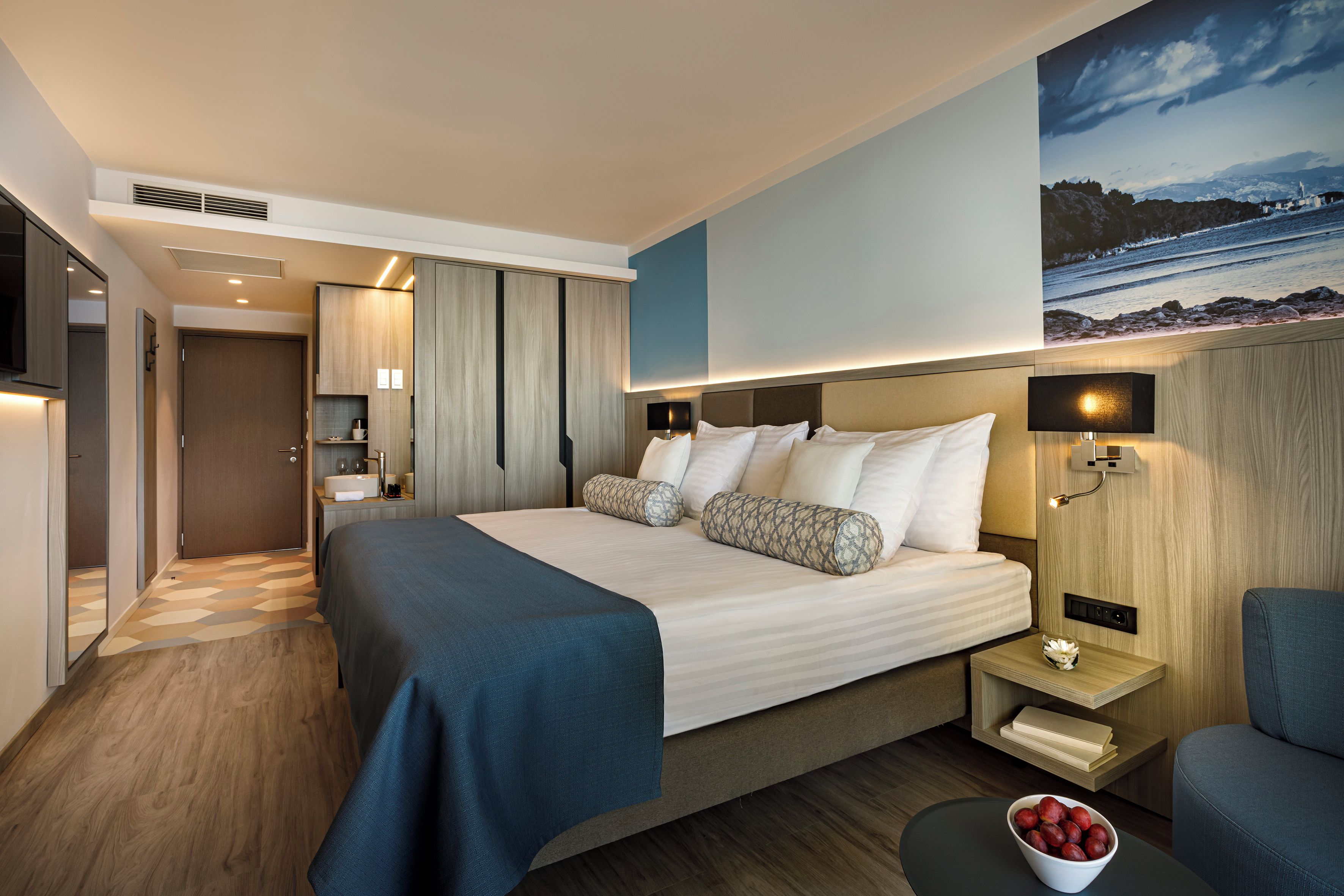 A modern hotel room with a large bed, bedside tables, a flat-screen TV, and a view of a coastal landscape.