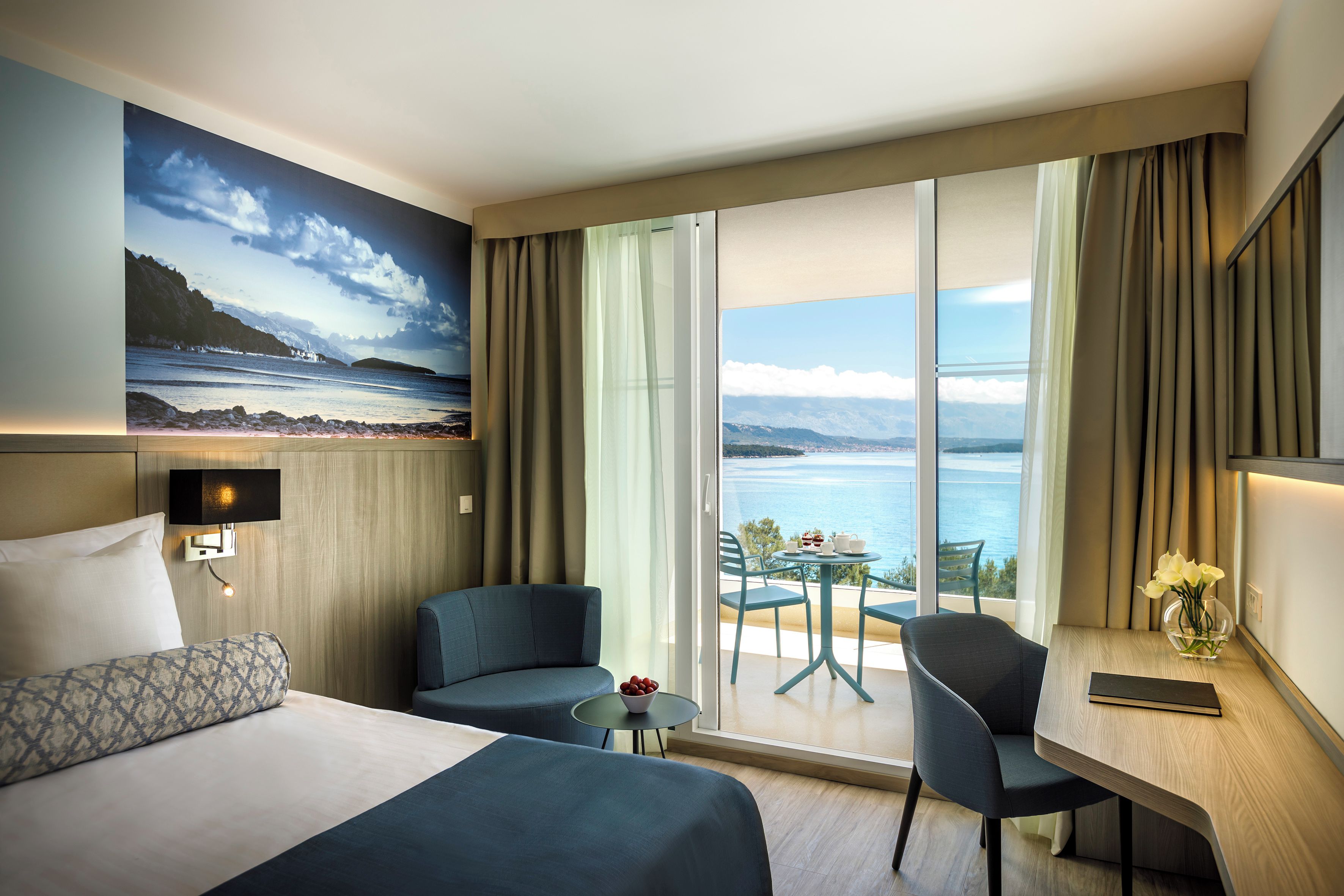 A hotel room with a bed, two armchairs, a small table, a desk, a painting, and a view of the sea.