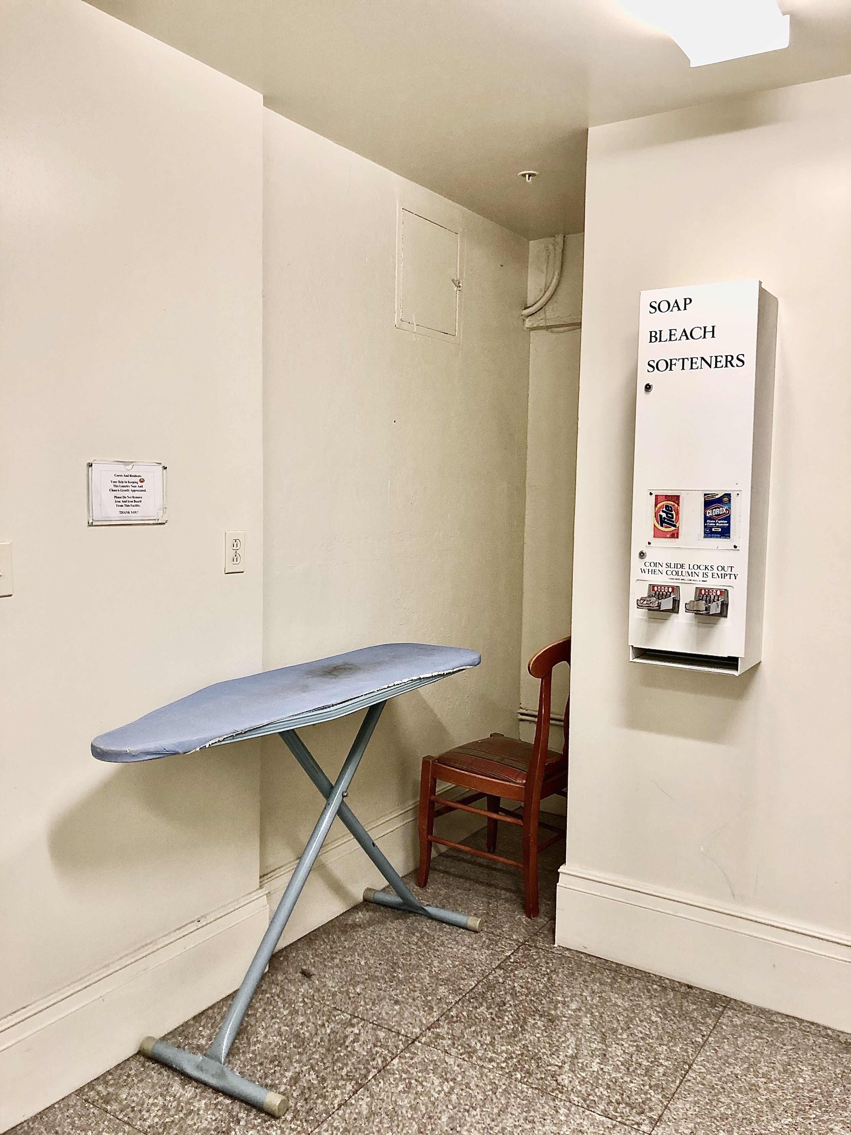 A small room with a portable ironing board, a chair, and a wall-mounted soap and bleach dispenser.