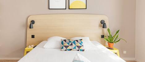A neatly made bed with a white comforter, a geometric patterned pillow, and a folded towel with a welcome card. There are two framed artworks above the bed, a nightstand with a phone and a plant on the right side.