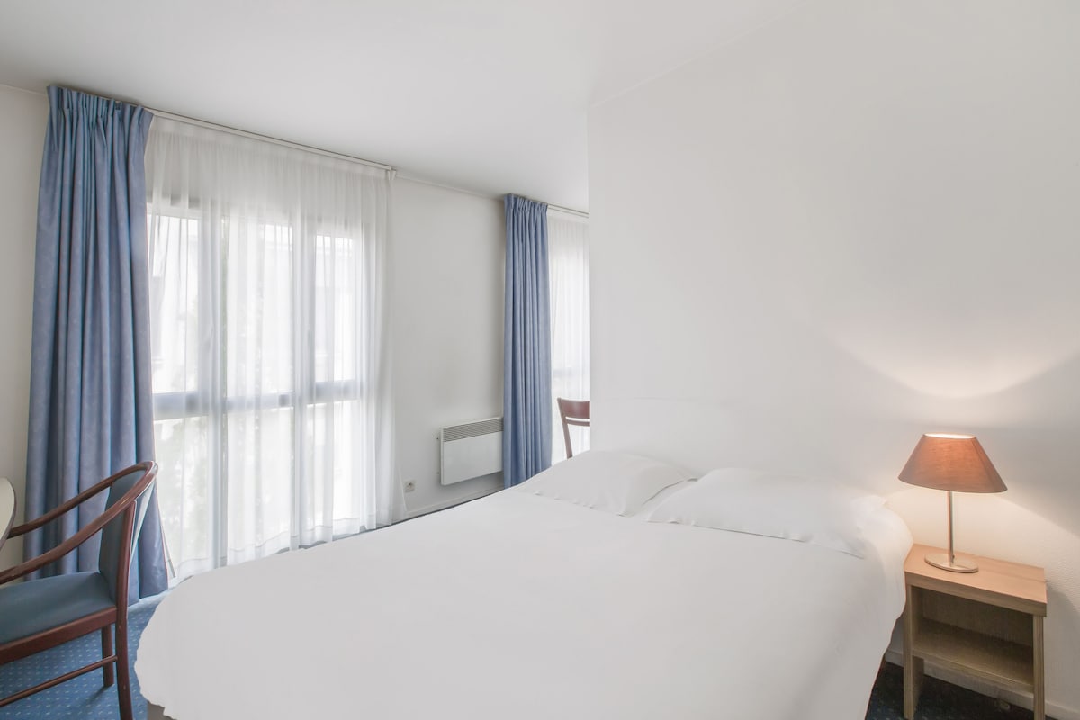A hotel room with a large bed, a bedside table with a lamp, a chair, and a window with curtains.