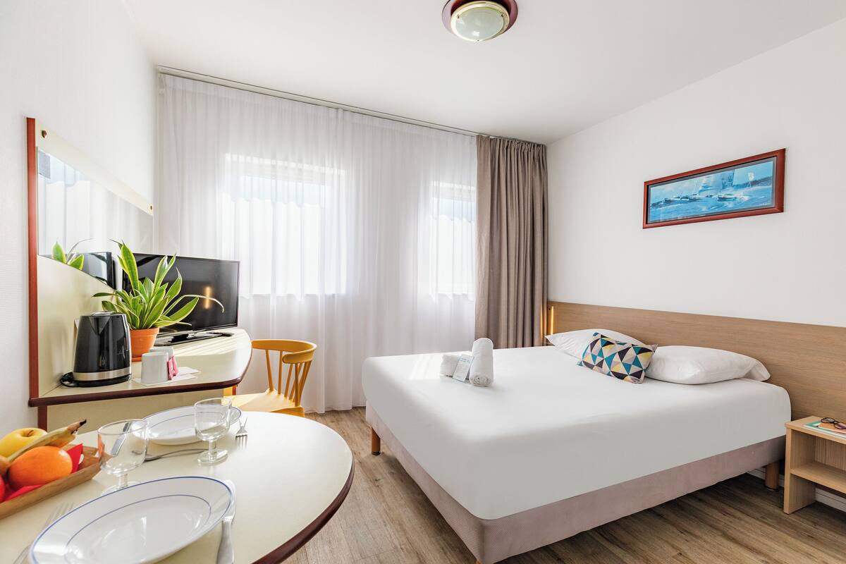 Une chambre d’hôtel avec un lit, un bureau avec une télévision, une table à manger et une fenêtre avec des rideaux.