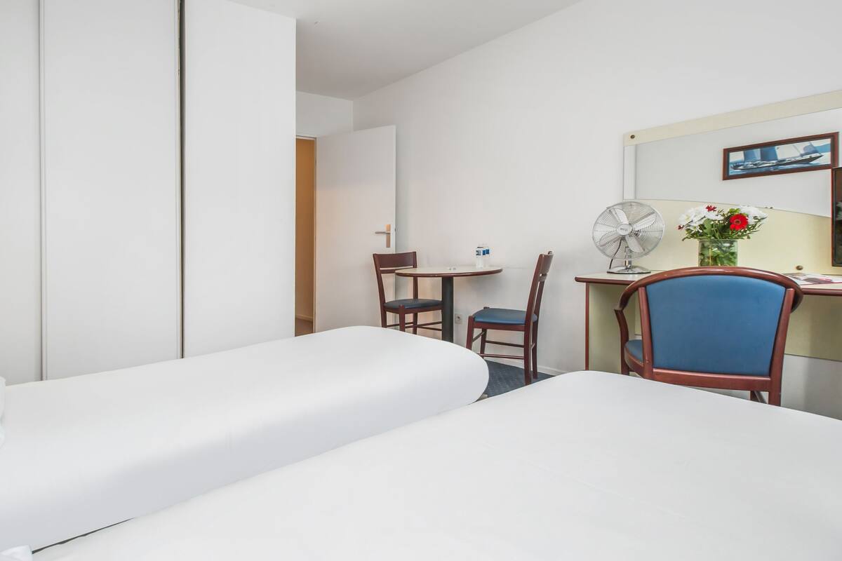 Une chambre d’hôtel avec deux lits, un bureau, une chaise, un ventilateur et un vase avec des fleurs.