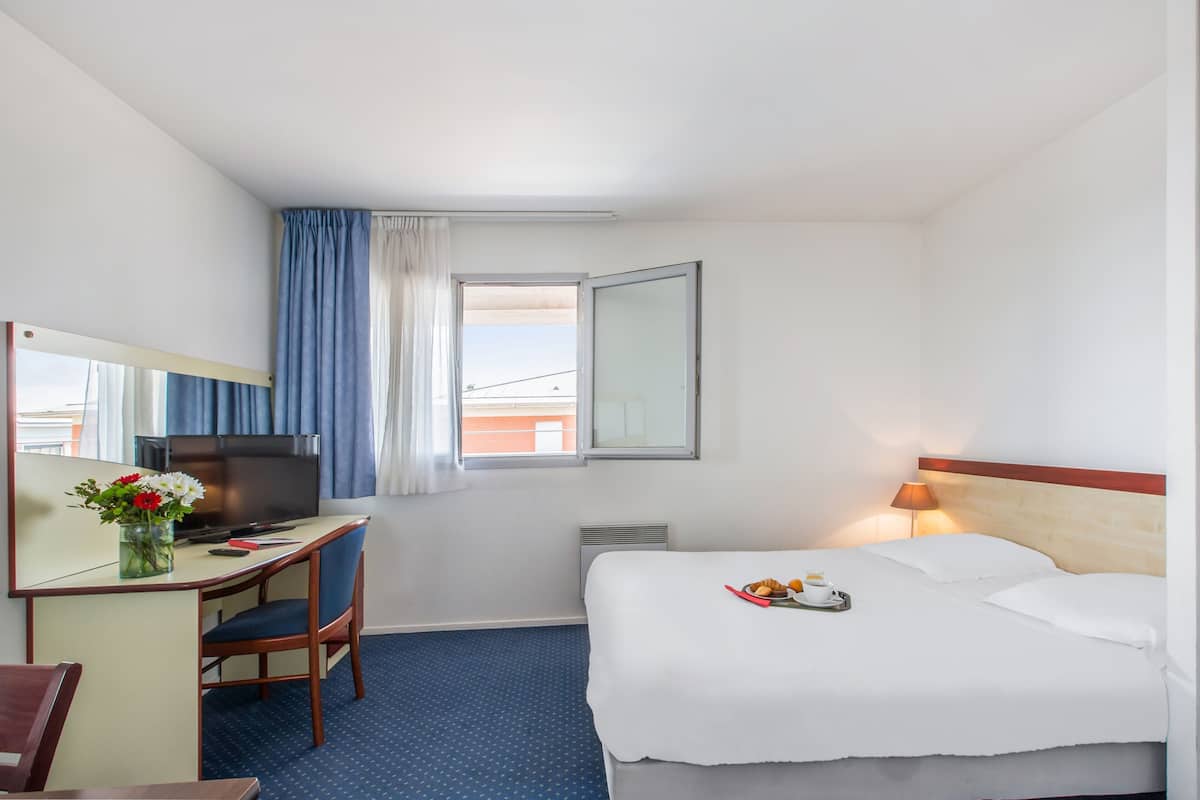 Une chambre d’hôtel comprenant un lit, un bureau avec une télévision, une chaise, une fenêtre avec des rideaux et une lampe.
