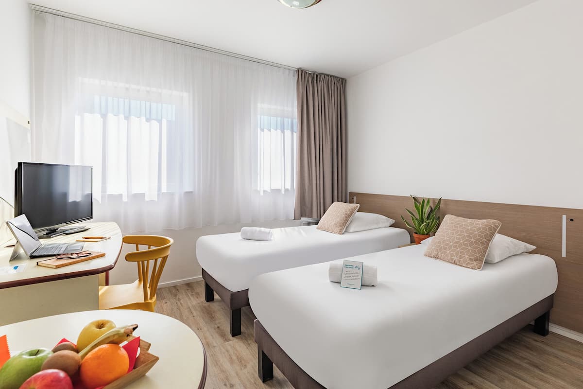Een hotelkamer met twee bedden, een bureau met een laptop, een schaal fruit en een kamerplant.