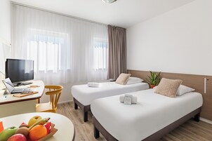 Une chambre d’hôtel avec deux lits, un bureau avec un ordinateur portable, un panier de fruits et une plante en pot.