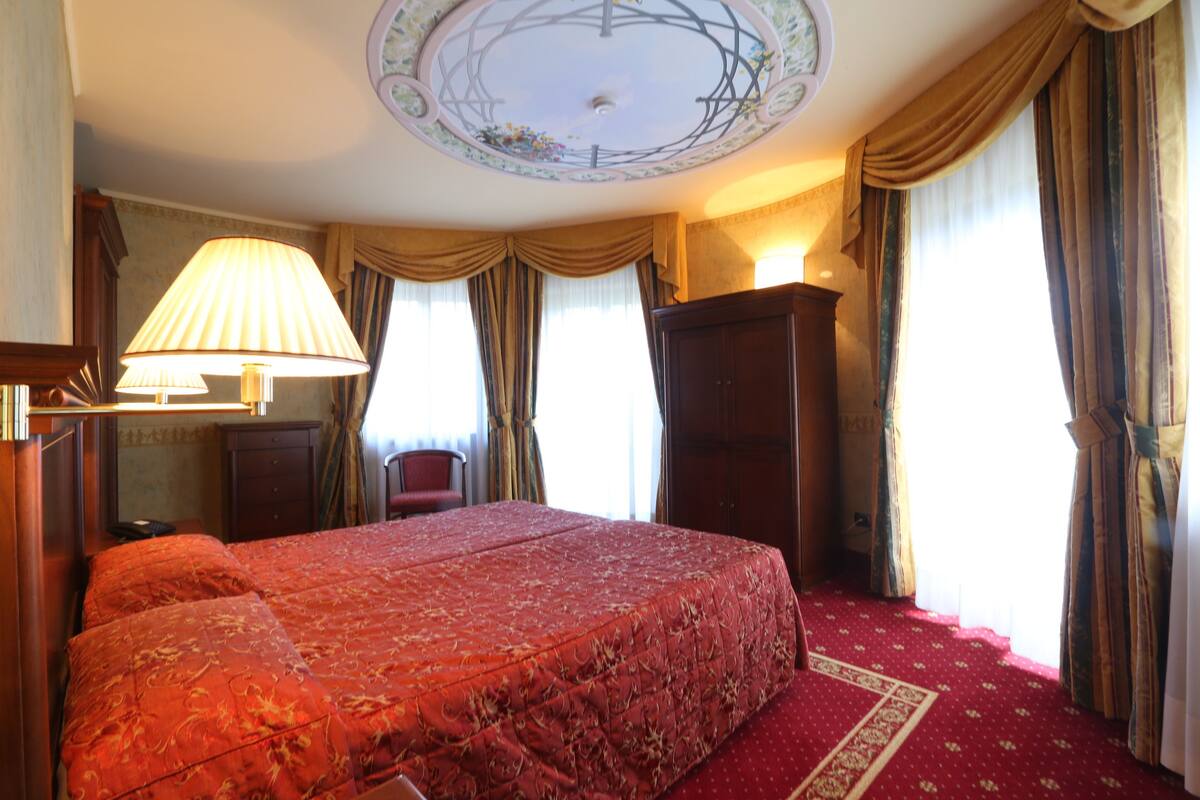 Habitación de hotel con una cama grande, un escritorio con lámpara, un armario y vistas al exterior a través de cortinas.