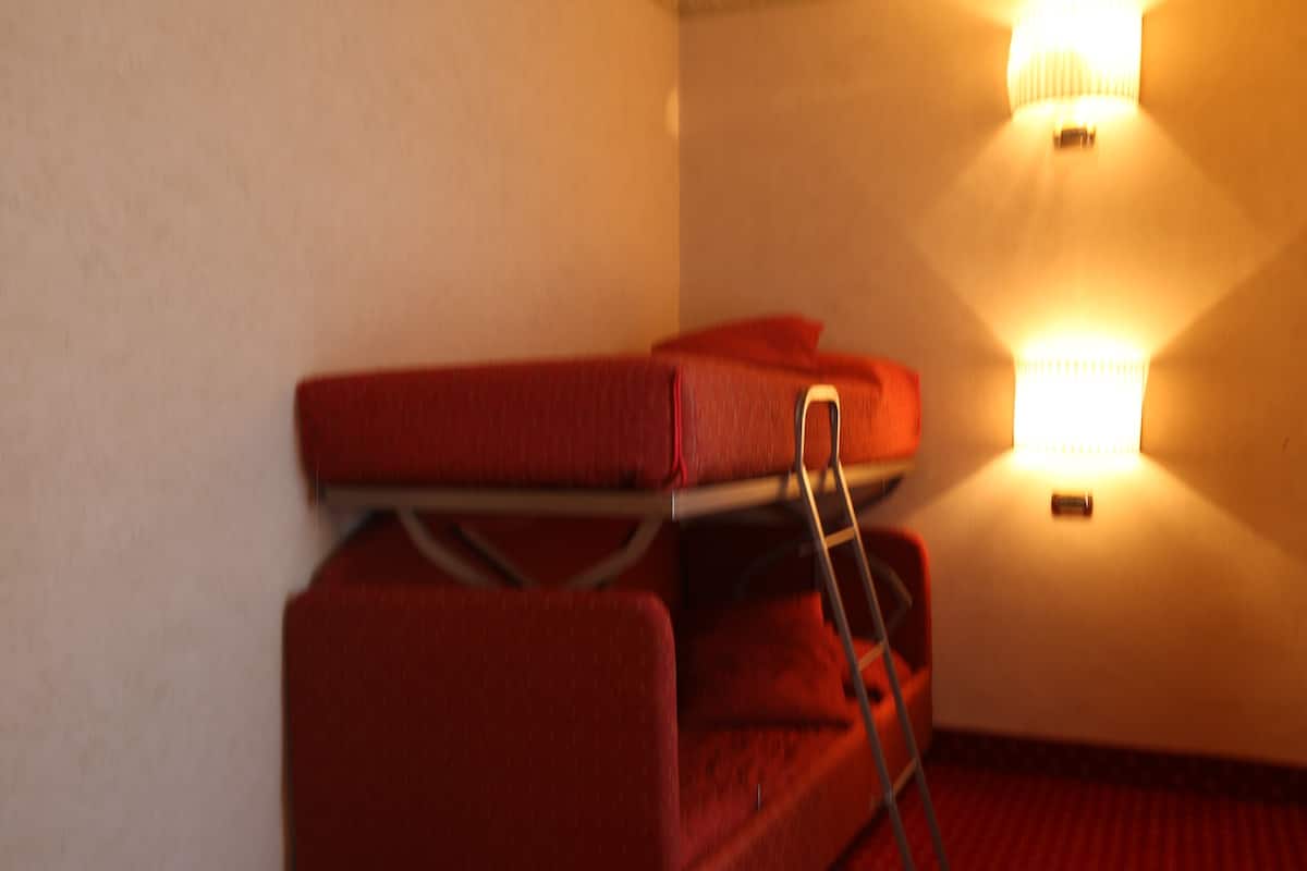Un rincón de una habitación con una maleta roja sobre un perchero metálico adosado a la pared, una silla roja y una lámpara fijada a la pared.
