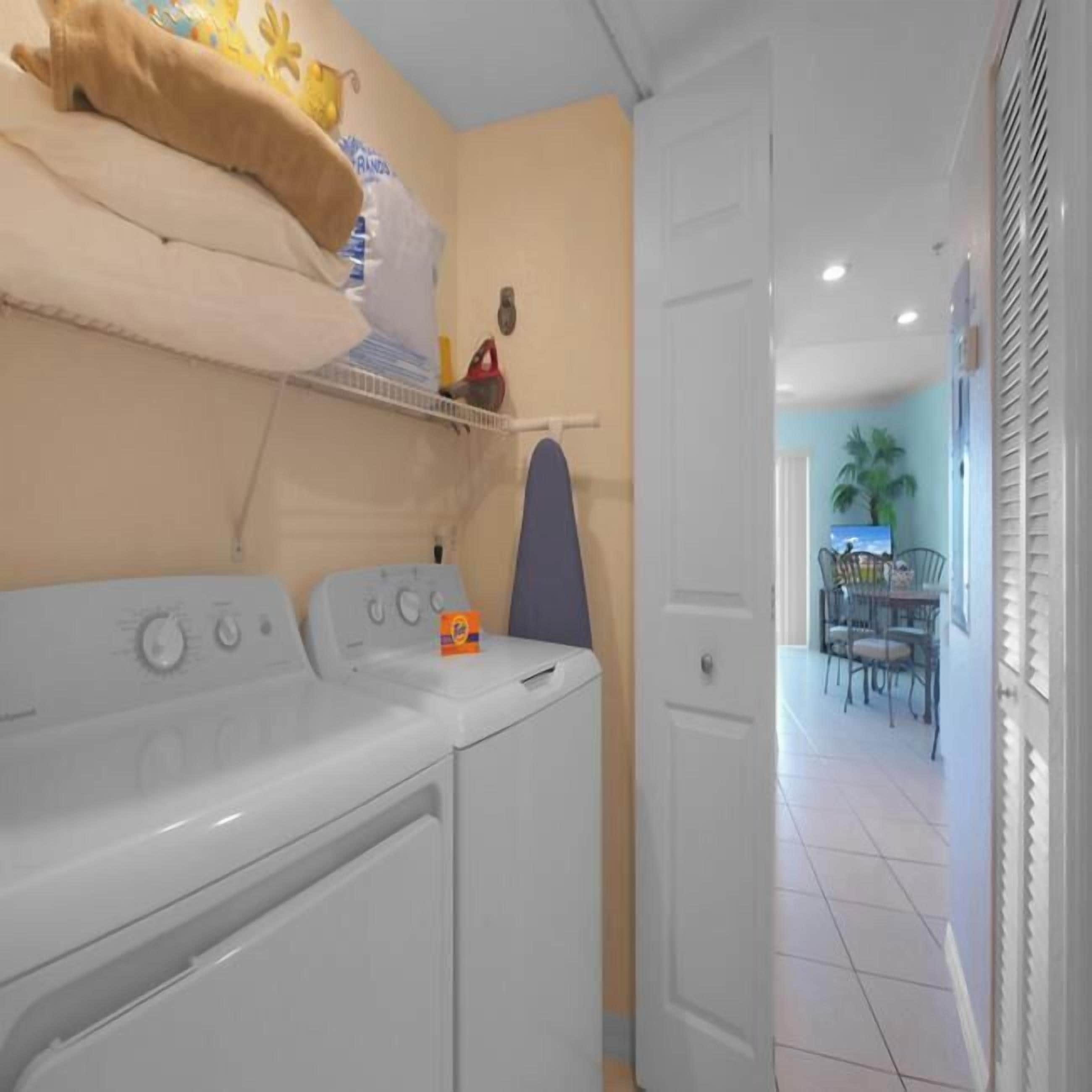 A laundry area with two washing machines, a shelf with towels and a small box, and a door leading to a dining area with a table and chairs.