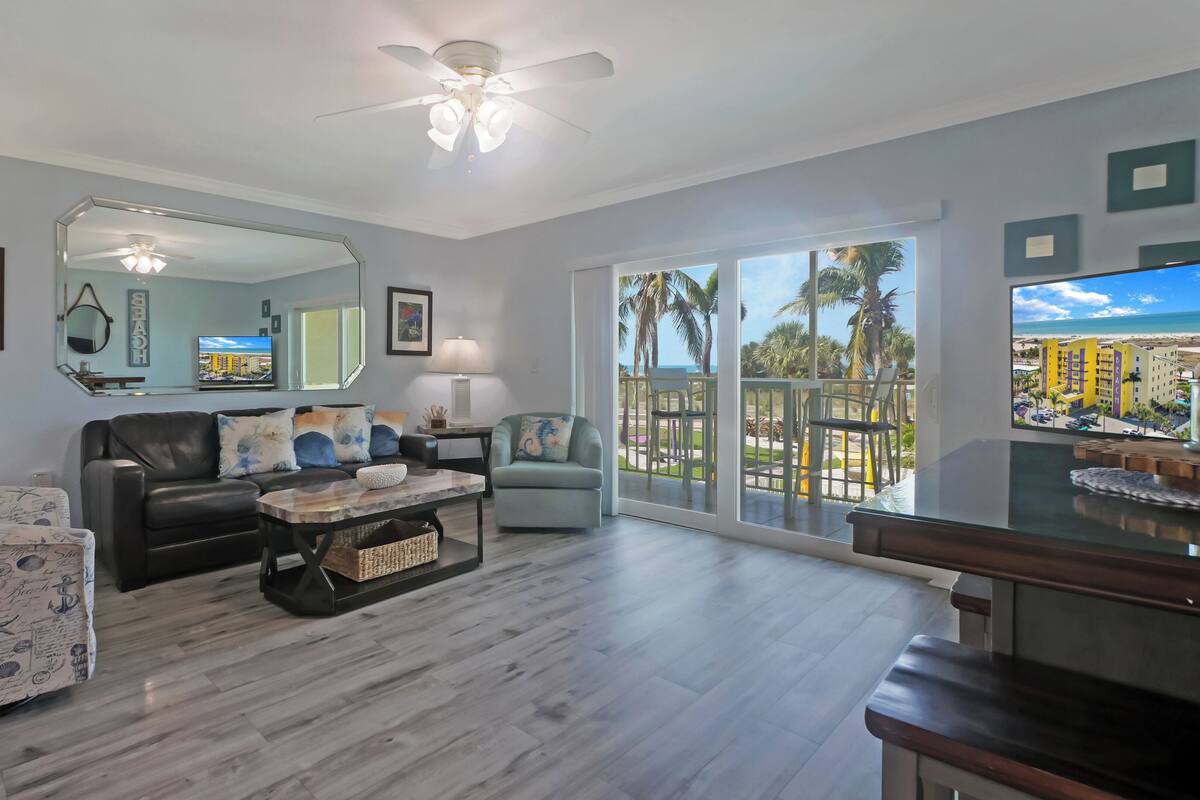 A spacious living room with a large mirror, a comfortable sofa, a coffee table, and a view of the beach through large windows.