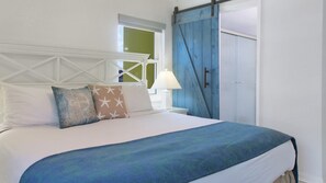 Una cama con sábanas blancas, una manta azul y almohadas estampadas con estrellas. Se ve una ventana con vistas a un paisaje verde y una puerta azul.
