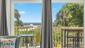 A balcony with a view of the beach, a table, a chair, and a decorative pillow.