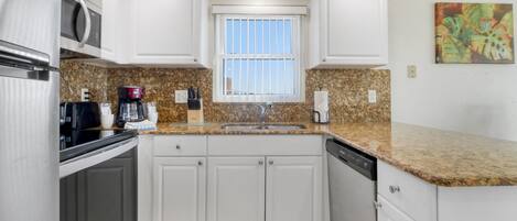A kitchen with white cabinets, a granite countertop, a stainless steel refrigerator, a coffee maker, and a window with blinds.