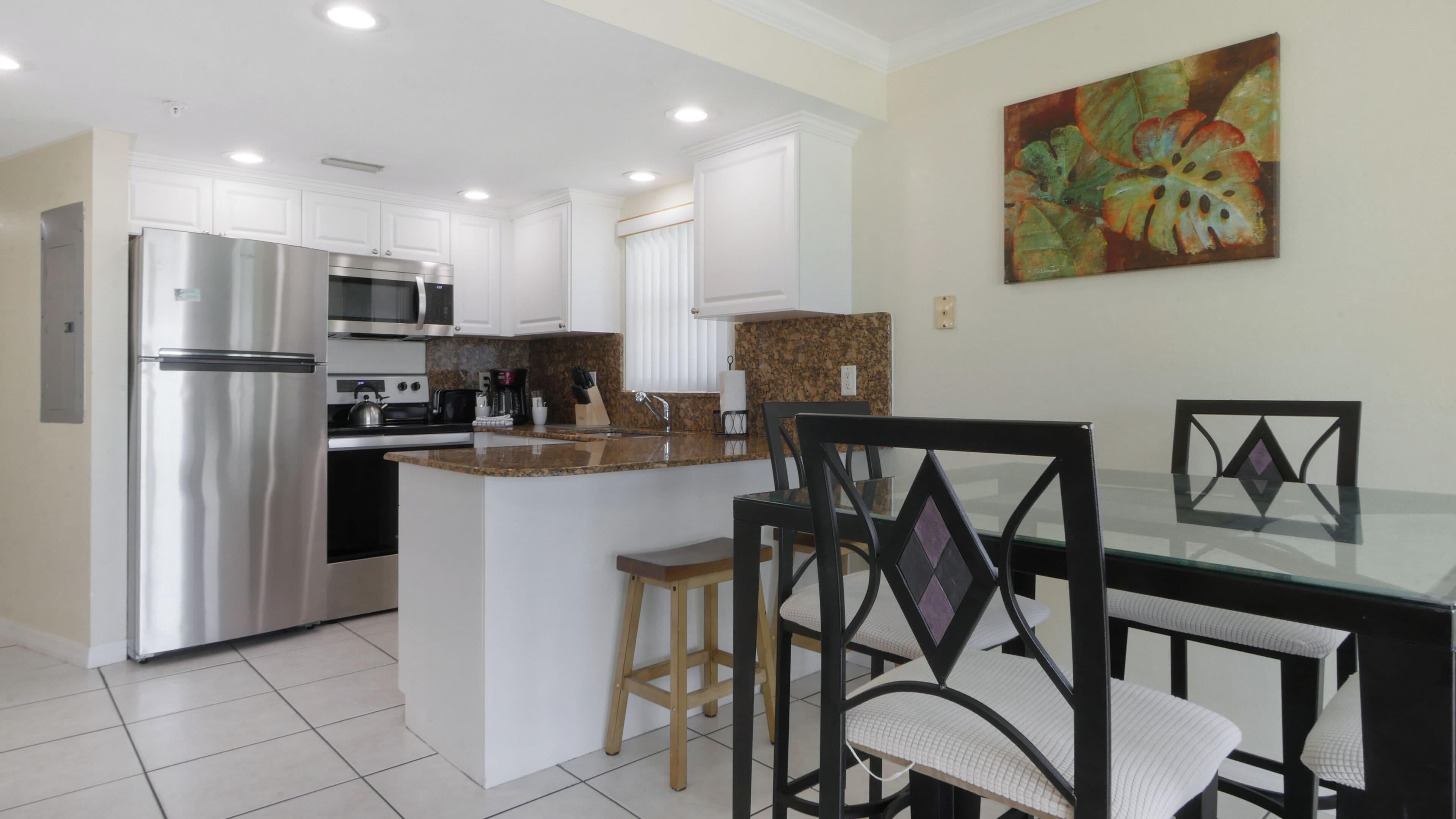 A modern kitchen with white cabinetry, stainless steel appliances, and a granite countertop. There is a dining area with a glass table and black chairs.