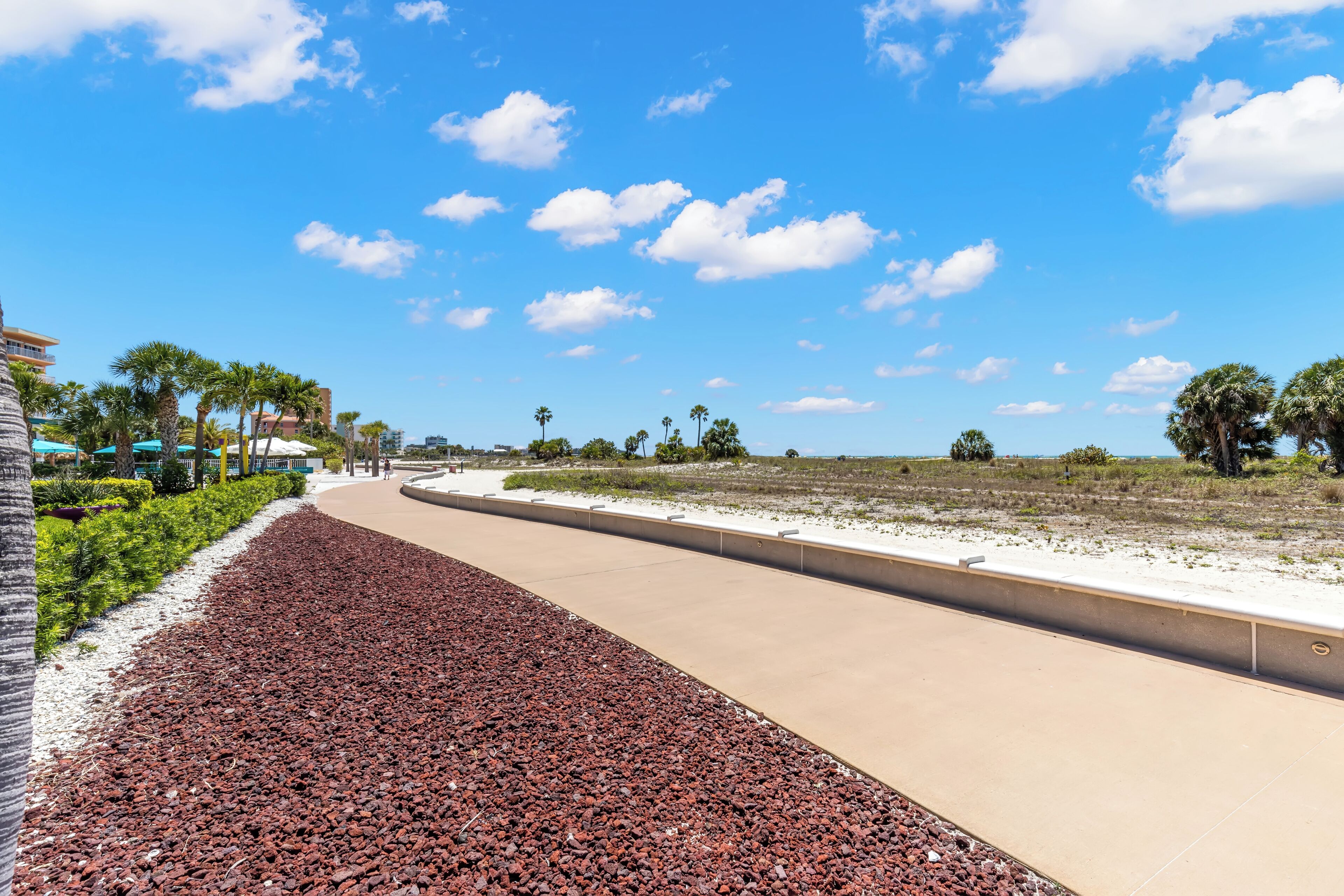 A paved walkway with red gravel, palm trees, and a clear blue sky.