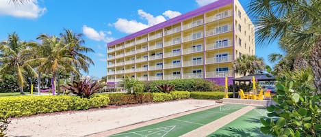Ein mehrstöckiges Hotel mit Balkonen, einem grünen Außen-Shuffleboard-Spielfeld und Palmen.