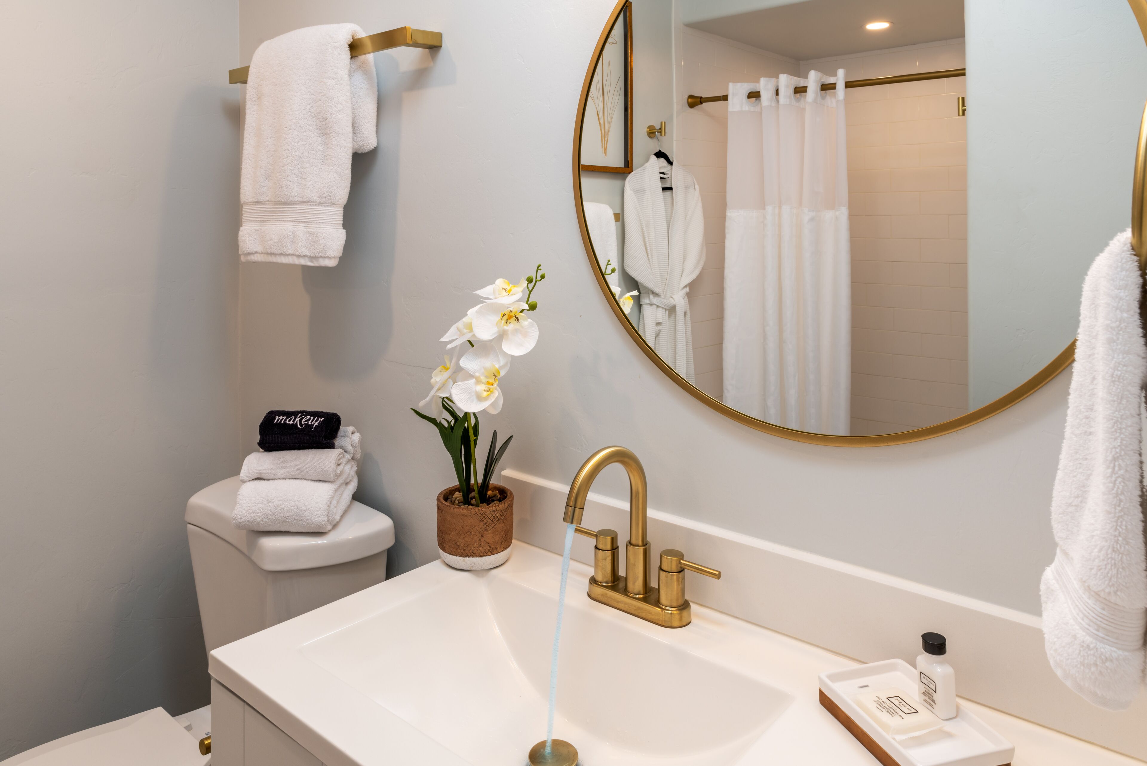 A modern bathroom with a white sink, gold faucets, a round mirror, and a potted orchid.