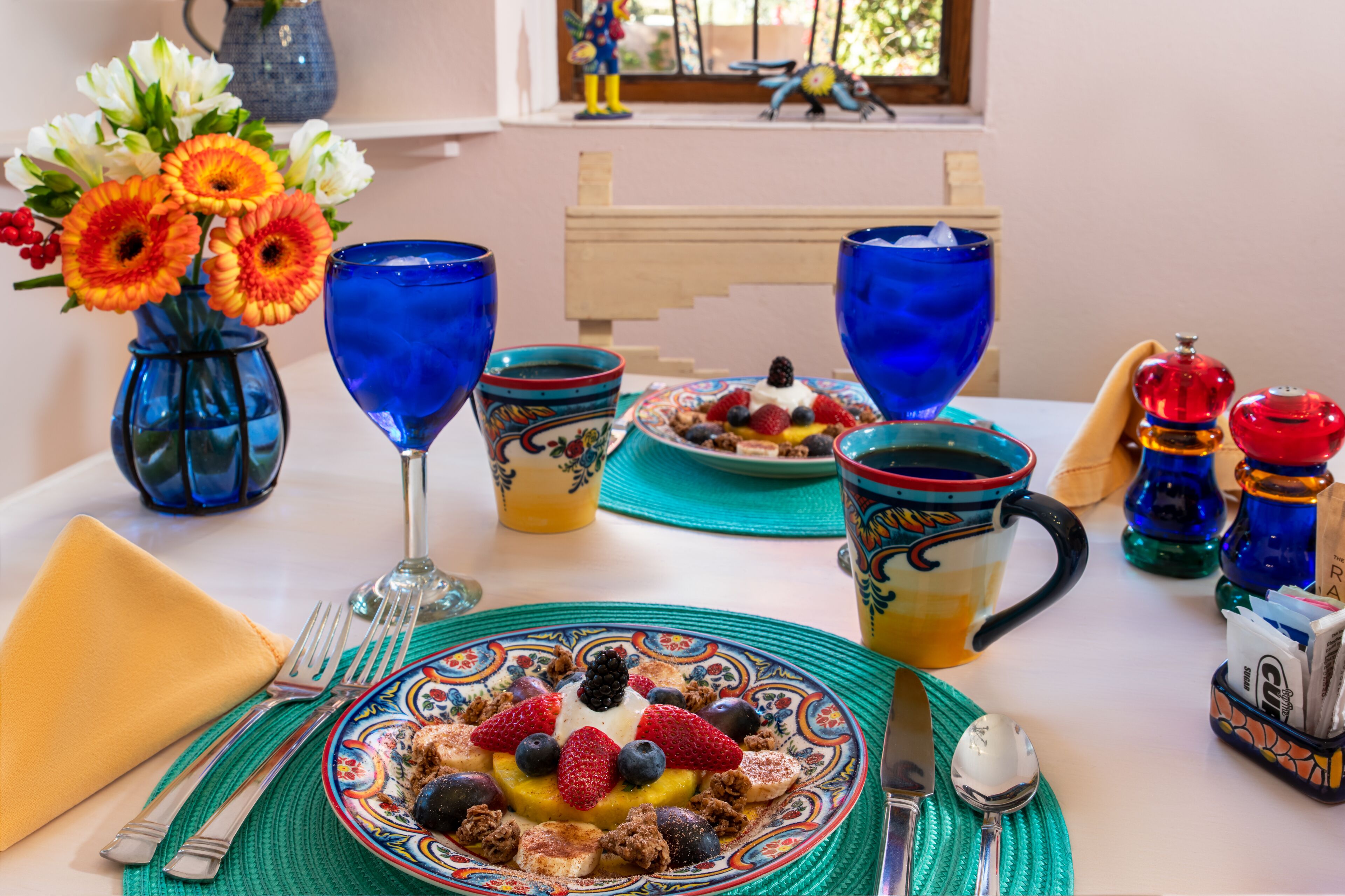 Una mesa de desayuno con una fuente de fruta, un florero y cristalería azul.