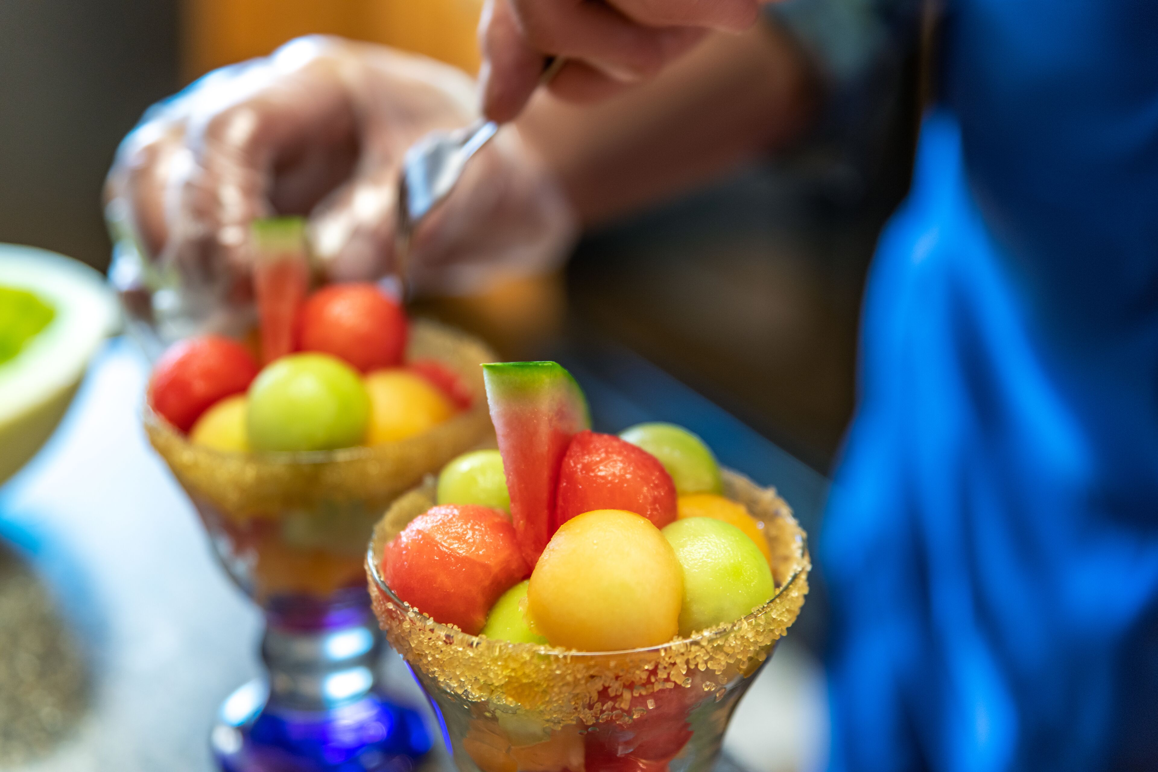 Primer plano de un plato de frutas con borde azucarado, una cuchara y una persona al fondo.