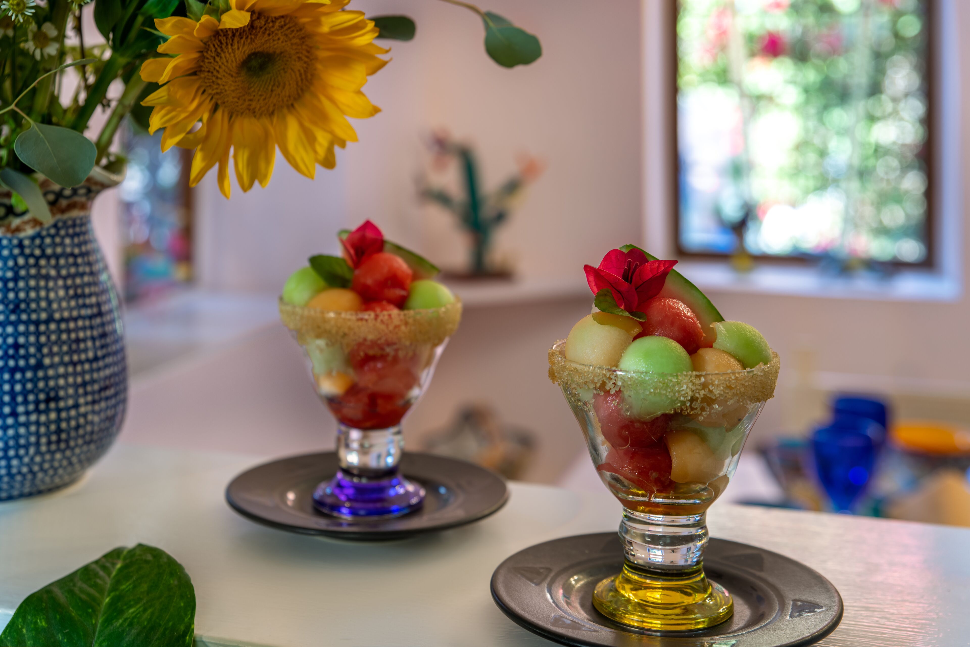 Two dessert glasses on a table with a vase of sunflowers and a decorative cactus in the background.