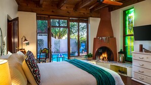 A bedroom with a large bed, a fireplace, a TV, and a view of a pool and garden. - Adobe Rose Inn B&B (Tucson)