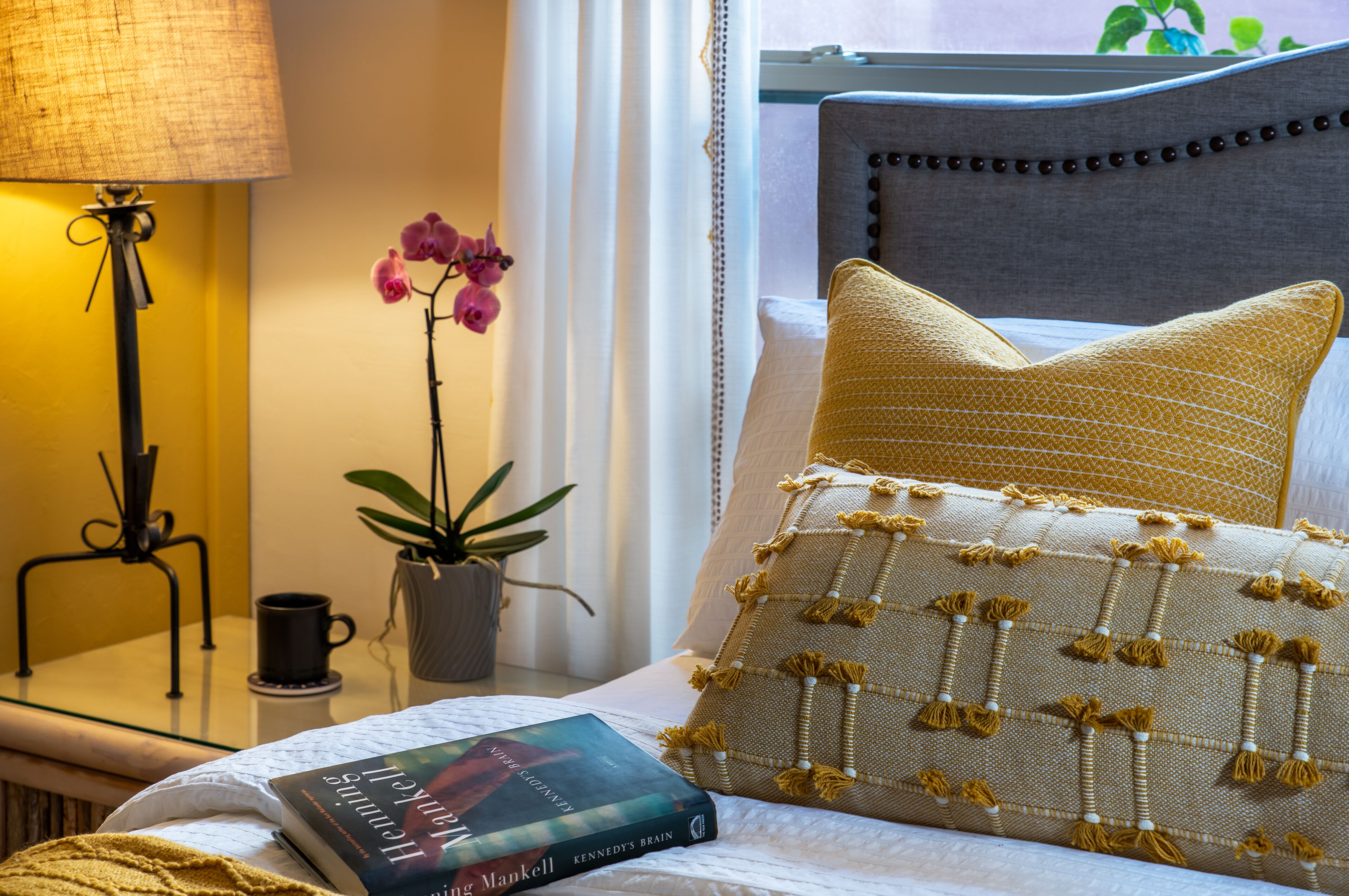 Ein ordentlich bezogenes Bett mit einem gemusterten Kopfkissen, ein Buch mit dem Titel "Kennedy's Brain", eine Lampe, ein Blumentopf und ein Fenster mit Vorhängen.
