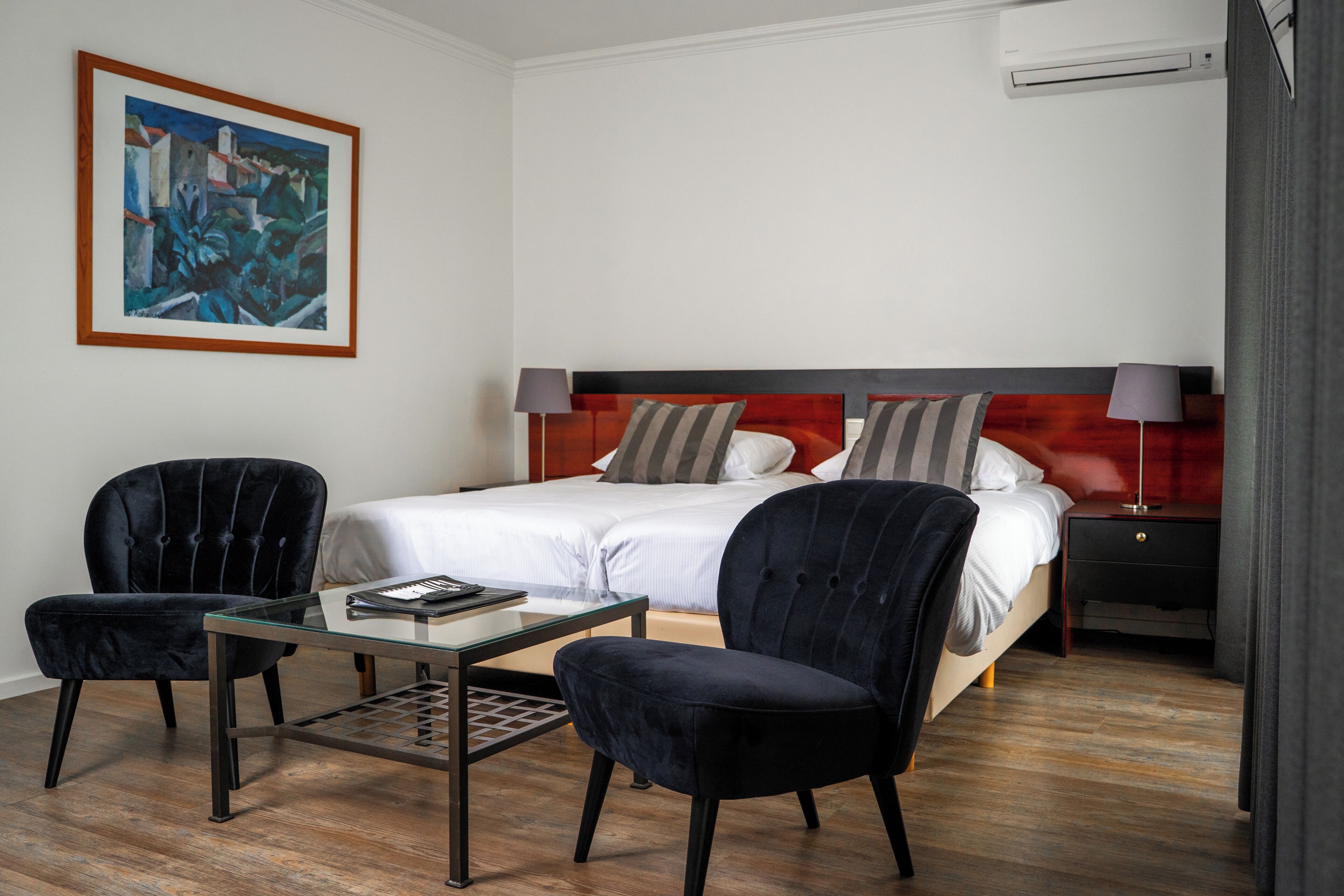 A hotel room with a bed, two chairs, a small table, a painting on the wall, and an air conditioning unit.