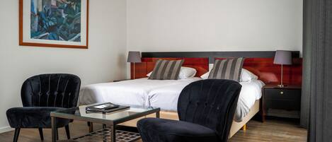 A hotel room with a bed, two chairs, a small table, a painting on the wall, and an air conditioning unit.