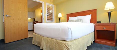 A hotel room with a large bed, a wooden nightstand with a lamp, a door leading to a bathroom, and a small table with a lamp.