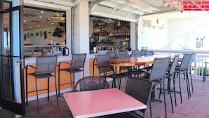 An outdoor dining area with wicker chairs and a wooden table, a bar with shelves of bottles, and a view of the street. - Ocean Sands Resort, Oceanfront, Virginia Beach by Vacatia (Virginia Beach)