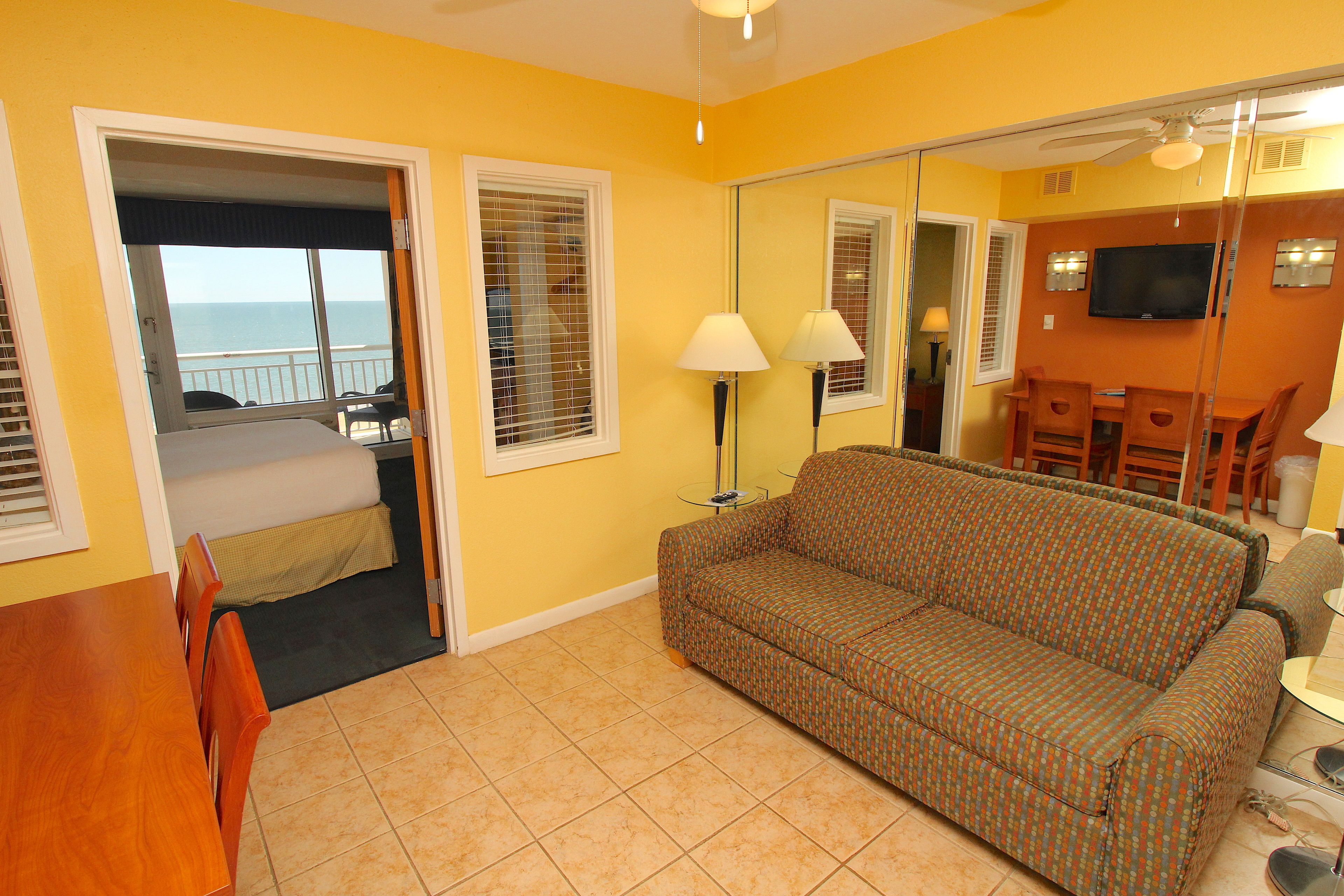 A room with a sofa, a dining table, and chairs. There is a television on the wall and a window with blinds.