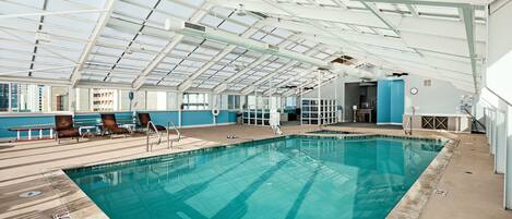 An indoor swimming pool with clear glass ceilings, lounge chairs, and a view of the city.