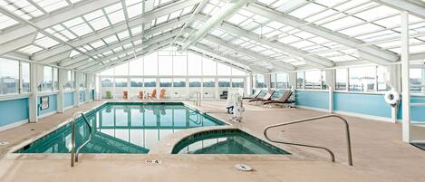 Una piscina interior con techos de vidrio transparente, sillas de descanso y vistas a la ciudad.