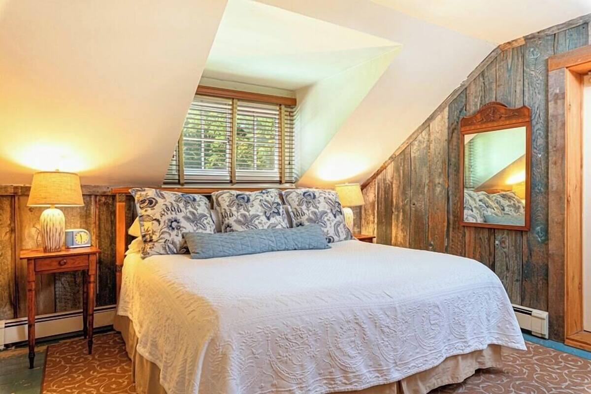 A bedroom with a large bed, a wooden headboard, a nightstand with a lamp, and a window with blinds.