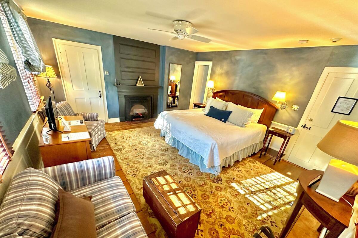 A hotel room with a large bed, a sofa, a wooden chest, a fireplace, and a ceiling fan.