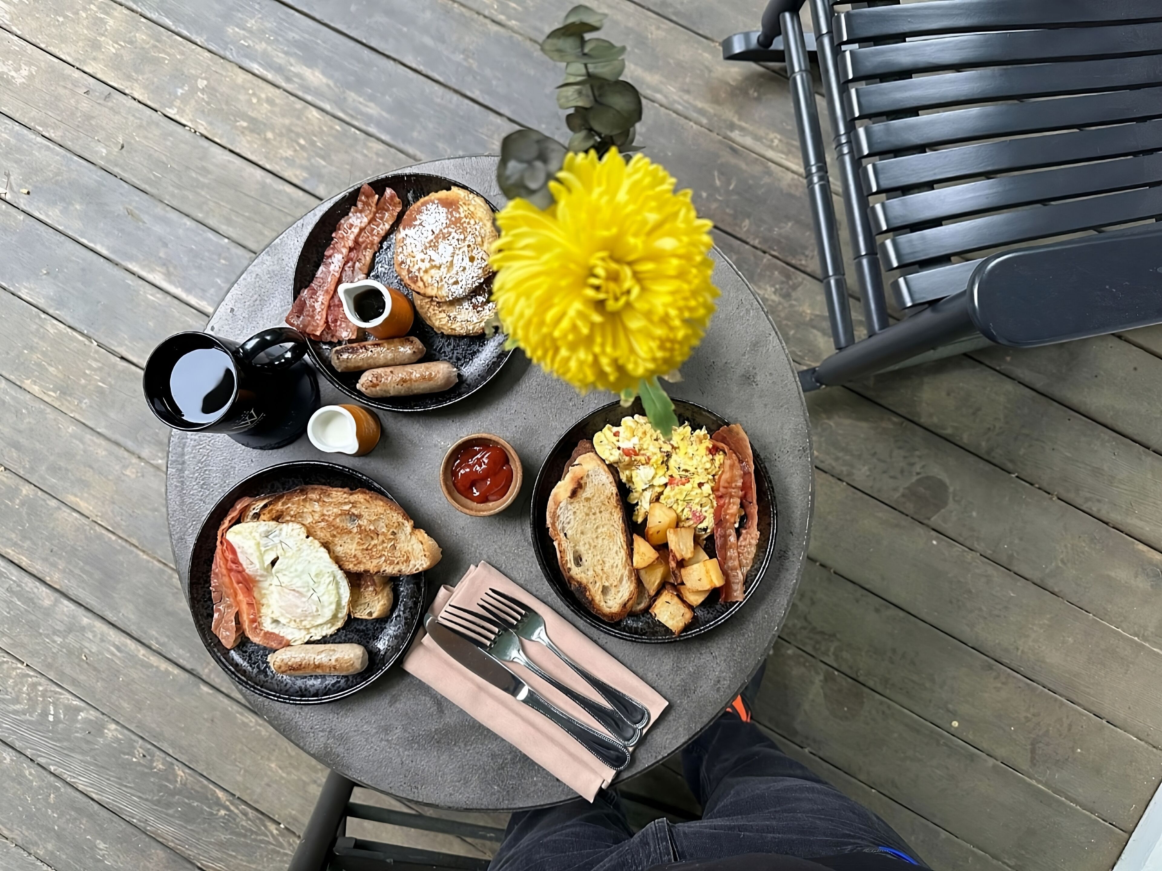 Et rundt bord med morgenmadsprodukter, blandt andet ristet brød, æg, pølser og en kaffekop.