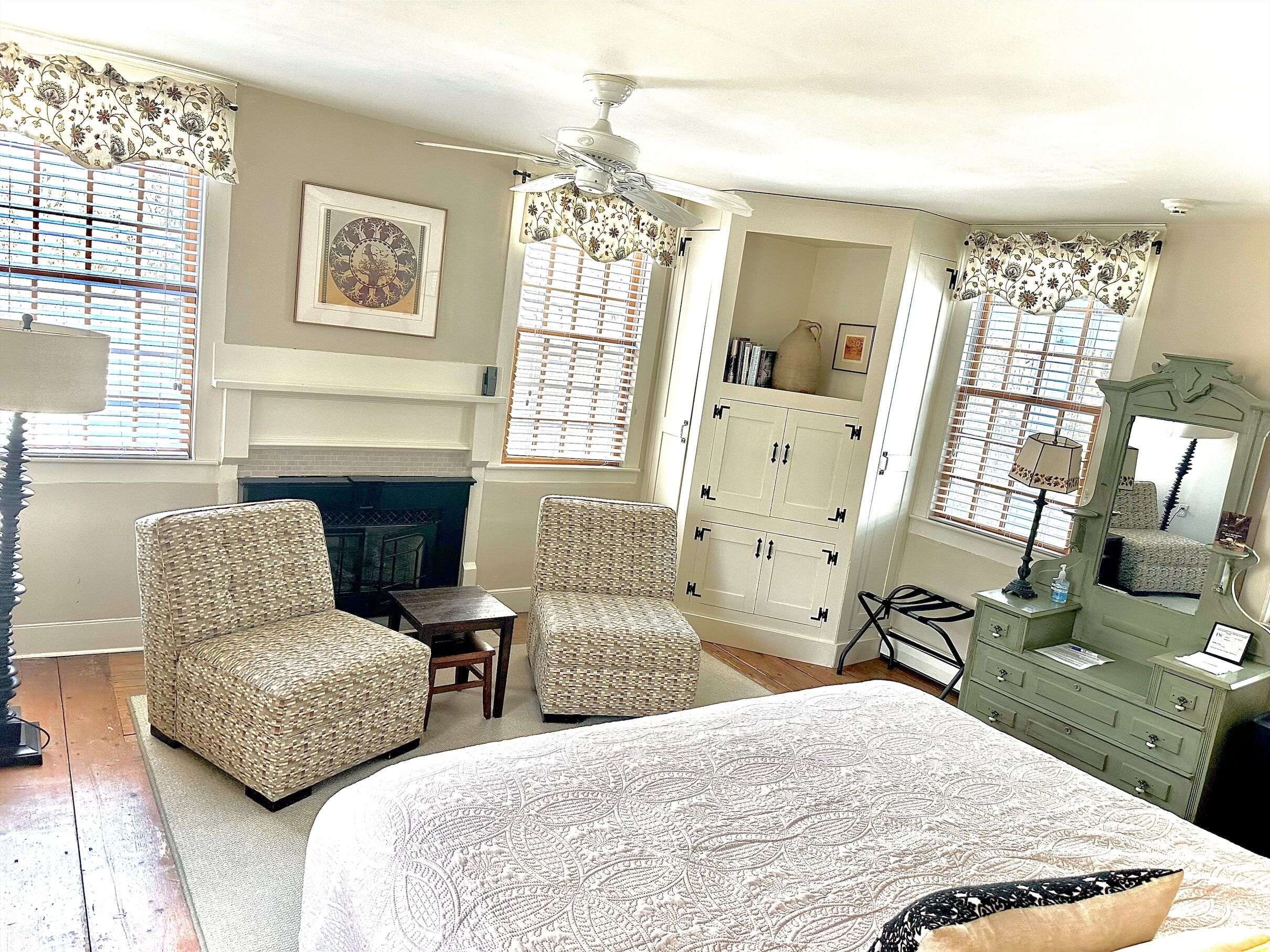 A bedroom with a large bed, two wicker chairs, a dresser, a mirror, and a fireplace.