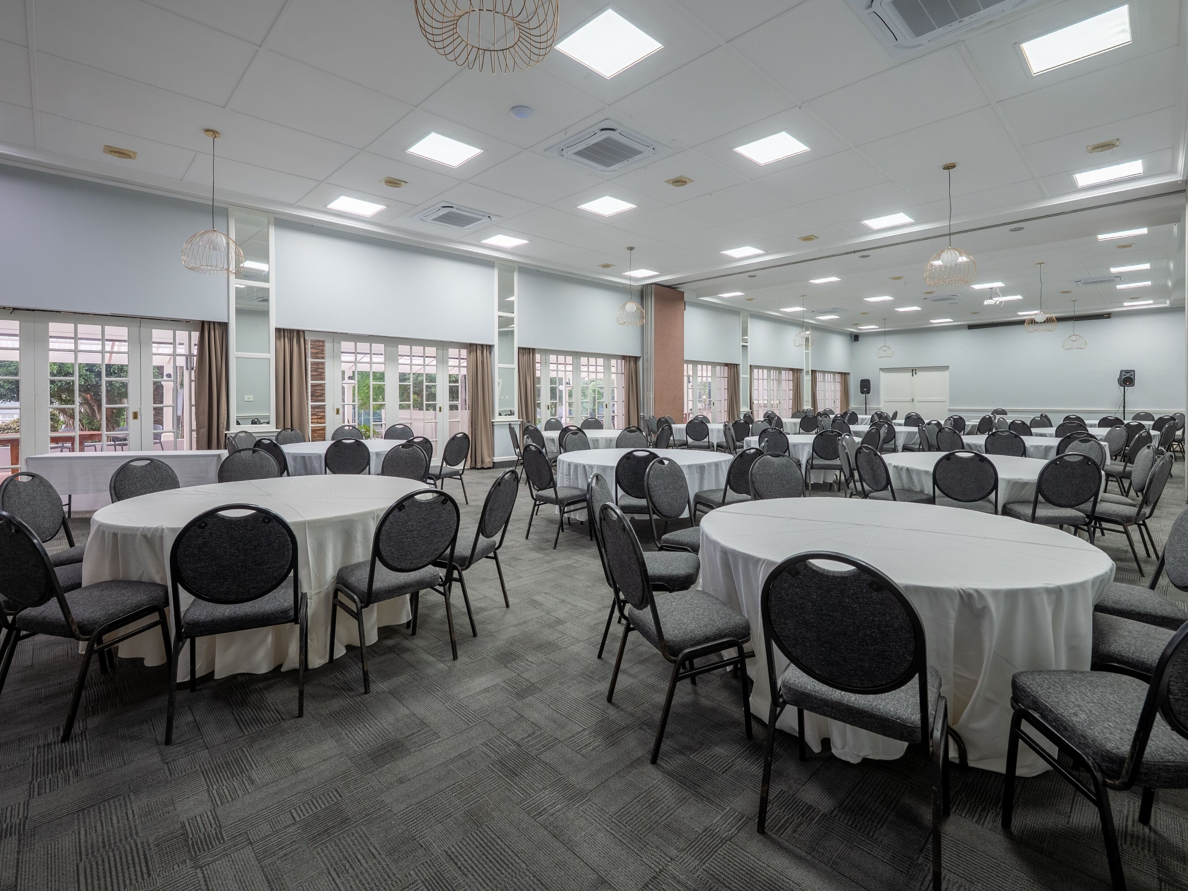 A conference room with multiple round tables and chairs, large windows, and ceiling lights.