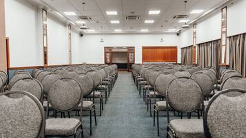 Uma sala de conferência com fileiras de cadeiras cinzas, uma porta de madeira na frente e um teto branco com luzes fluorescentes.
