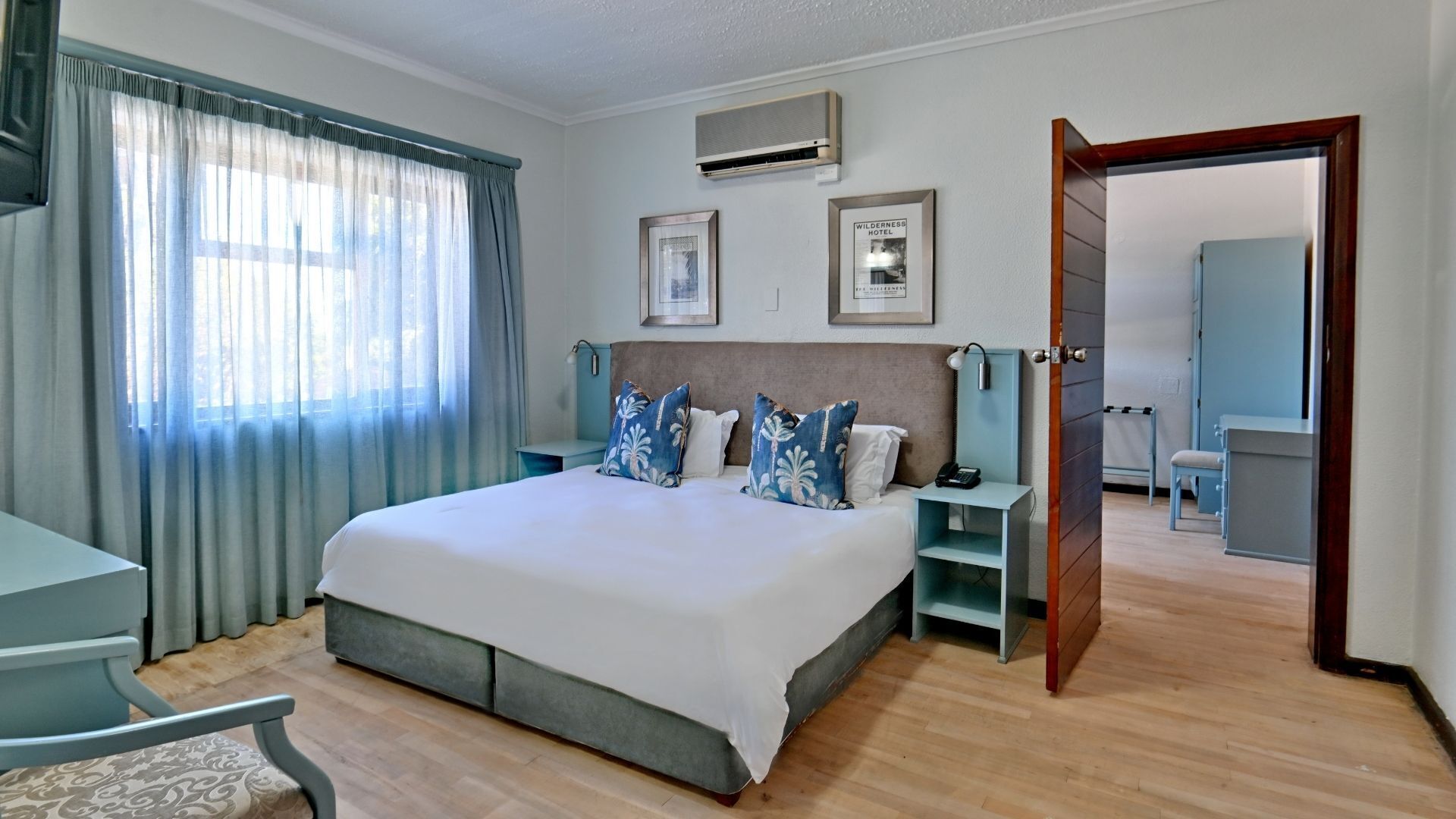 A hotel room with a bed, bedside tables, a desk, and a chair. There is a window with curtains, a door leading to another room, and a framed picture on the wall.