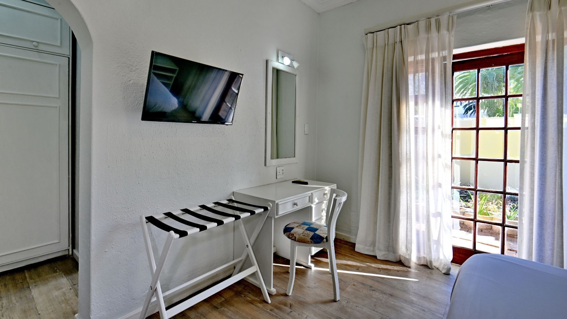 A room with a white desk, a mirror, a TV mounted on the wall, and a window with curtains.
