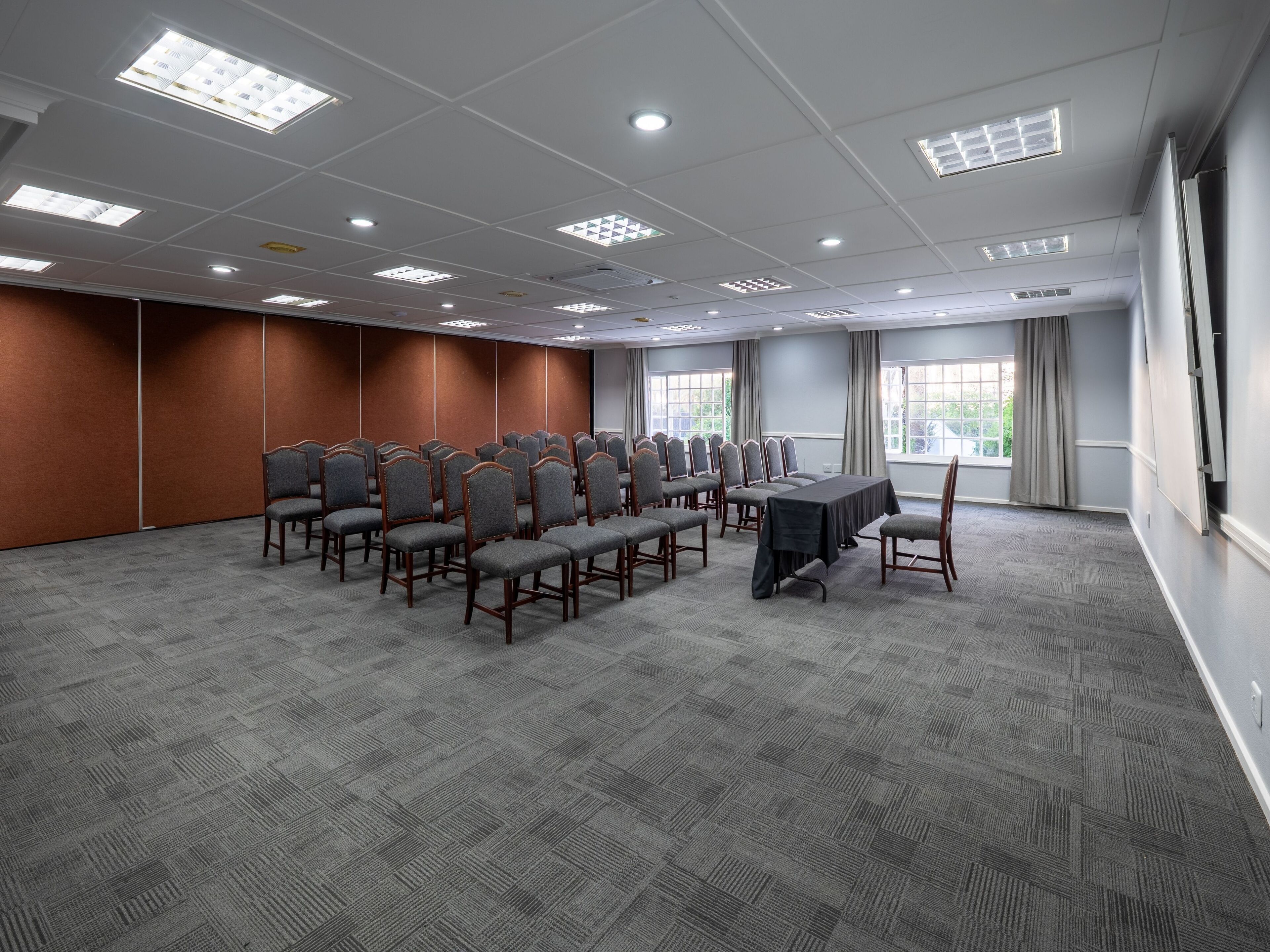 Un salón de conferencias con filas de sillas, una larga mesa y amplias ventanas con cortinas.