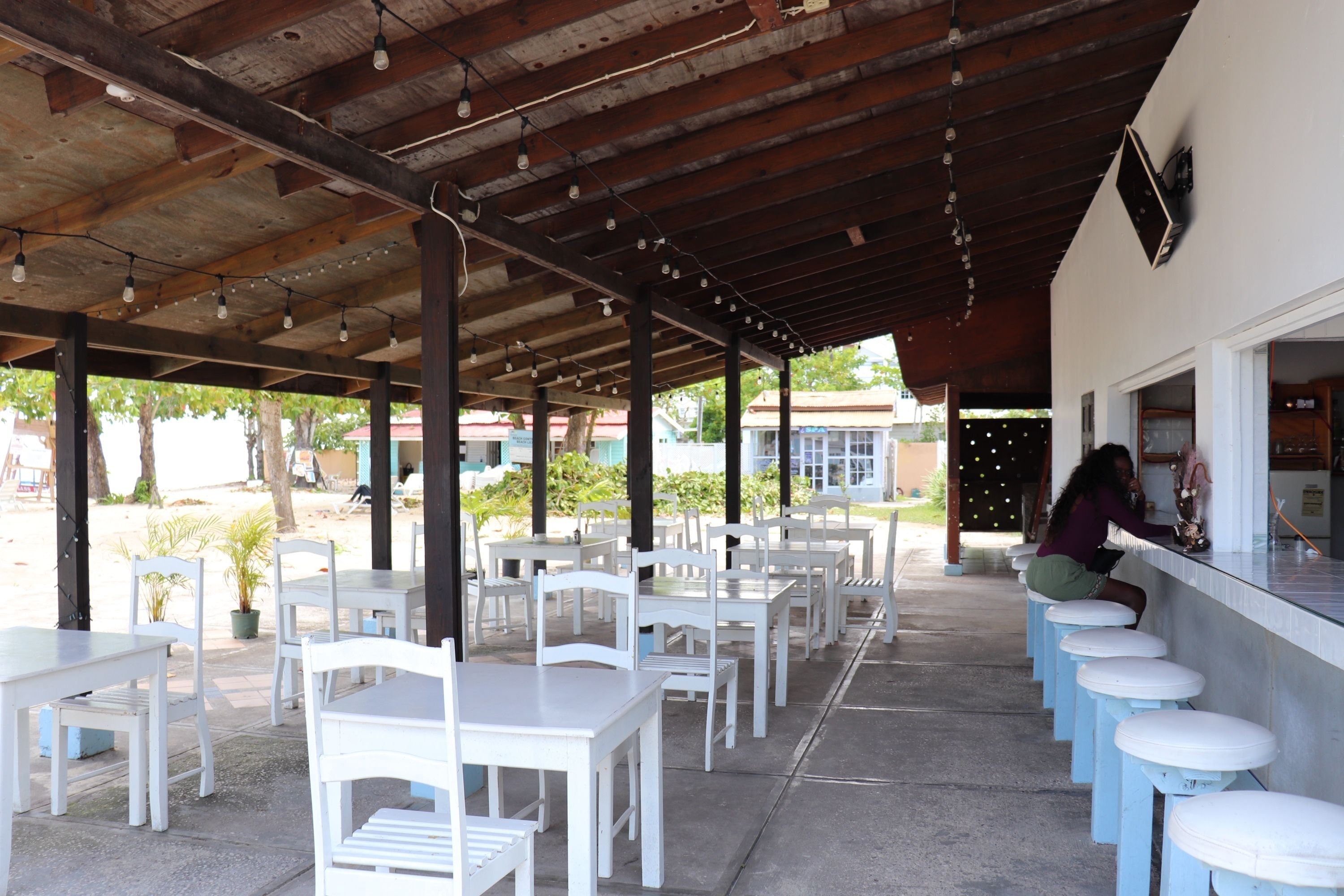 An outdoor cafe with white tables and chairs, a bar area with stools, and a covered ceiling with hanging lights.